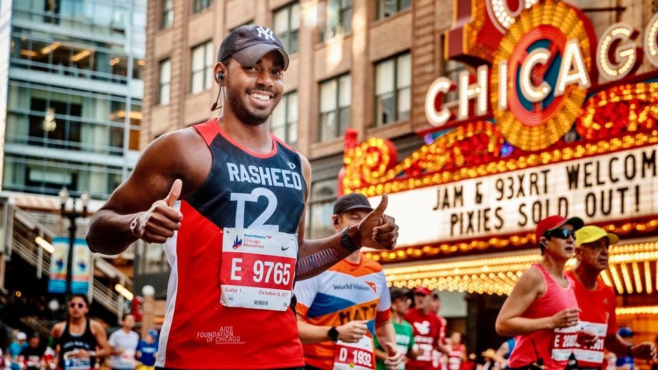 Rasheed Gonga running the 2017 Chicago Marathon in an AIDS Foundation of Chicago singlet, giving a thumbs up under the Chicago Theatre marquee